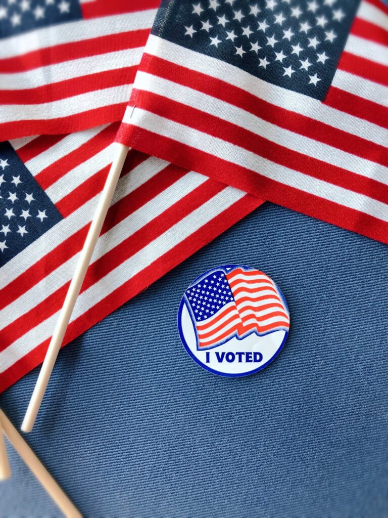 USA flags & an “I Voted” sticker overhead on a blue background. Voting is a citizen’s right & duty